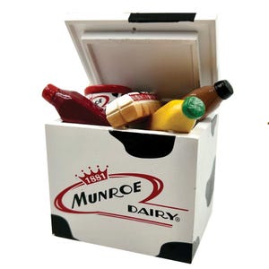 Puede incluir: Un mini refrigerador blanco con un diseño de vaca en blanco y negro. El refrigerador tiene un logotipo rojo y blanco que dice "Munroe Dairy" con el año "1881" sobre el nombre. El refrigerador está abierto y contiene artículos de comida en miniatura, incluyendo una botella de ketchup, una lata de sopa, una barra de pan, un plátano y una botella de jugo.