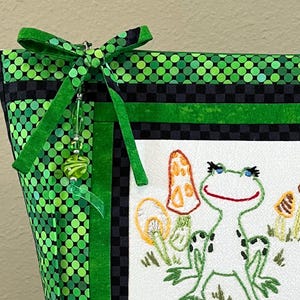 May include: A green and black fabric tote bag with a frog and mushroom embroidery design. The bag features a green polka dot pattern, a black and green checkered pattern, and a green bow with a beaded charm.