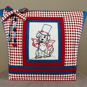 May include: A fabric tote bag with a red and white checkered pattern. The bag features an embroidered image of a dog wearing a top hat and bow tie. A blue bow and beaded charm adorn the side.