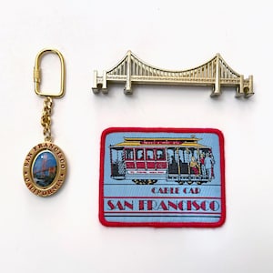 May include: A collection of San Francisco souvenirs, including a gold-colored keychain with an oval pendant, a gold-colored bridge pin, and a patch featuring a cable car. The patch has the words "Cable Car" and "San Francisco".