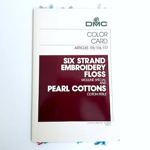 May include: A DMC color card for six strand embroidery floss and pearl cottons, featuring a white cover with a large maroon rectangle, black text, and the DMC logo. Small colorful thread samples are visible at the top and bottom edges.