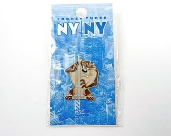 May include: A Looney Tunes pin featuring the Tasmanian Devil holding the Empire State Building. The pin is on a blue background with the text "NY NY" and "Looney Tunes". The pin is in a clear plastic package.