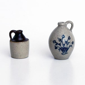 May include: Two miniature ceramic jugs. The smaller jug has a cylindrical body with a light tan base and a dark brown glazed top. The larger, rounded jug is light tan with a blue hand-painted floral design and a single side handle.