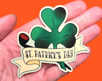May include: A St. Patrick's Day sticker showing a green shamrock with a black pipe and a banner reading "ST. PATRICK'S DAY". The sticker has a white border and is held in a hand against an orange background.