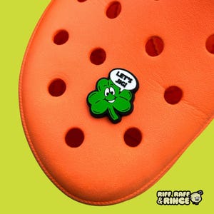 Op de afbeelding: Groene klavertje vier bedel met een glimlachend gezicht en een spraakballon met de tekst "Let's Jig!" De bedel is bevestigd aan een oranje schoen.