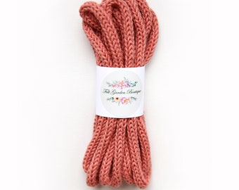 May include: A coiled length of dusty rose colored knitted cord secured with a white paper band. The band features a circular logo with a floral illustration and the text Felt Garden Boutique printed in a black script font across the center.