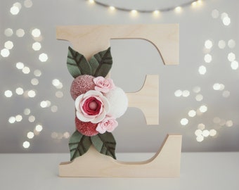 Puede incluir: Una letra E de madera grande decorada con un grupo de flores de fieltro y pompones. El arreglo incluye hojas de fieltro verde oscuro, un pompon blanco grande, un pompon rosa polvoriento y varias flores de fieltro en capas rosas y blancas.
