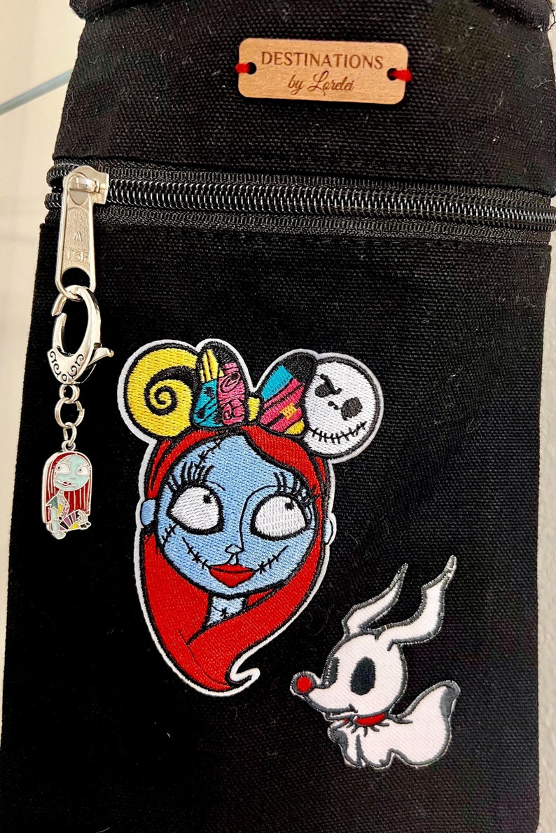 May include: Black canvas zippered pouch with a wooden tag that reads "Destinations by Lordel" and features embroidered patches of Sally from The Nightmare Before Christmas, Zero the ghost dog, and a Jack Skellington head.