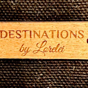 May include: A wooden tag with the text "DESTINATIONS by Loralei" in a cursive font. The tag is tied with red string.