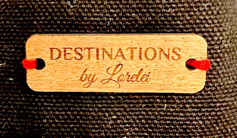 May include: A wooden tag with the text "DESTINATIONS by Loralei" in a cursive font. The tag is tied with red string.