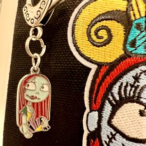May include: A silver metal keychain with a charm of Sally from the Nightmare Before Christmas. Sally is depicted with her signature red hair, green skin, and patchwork dress. The keychain is attached to a black fabric background with a large embroidered patch of Sally's face.
