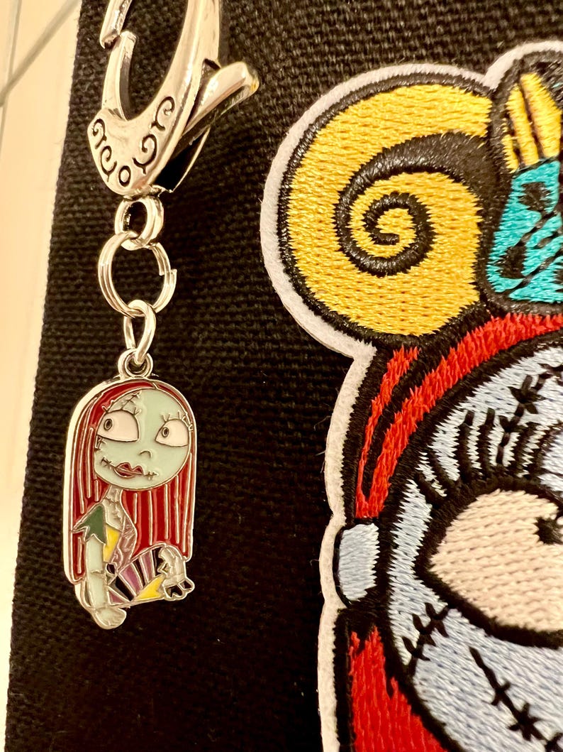 May include: A silver metal keychain with a charm of Sally from the Nightmare Before Christmas. Sally is depicted with her signature red hair, green skin, and patchwork dress. The keychain is attached to a black fabric background with a large embroidered patch of Sally's face.