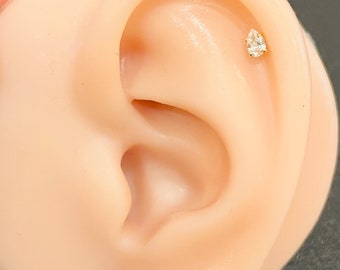 May include: A close-up of a person's ear with a small, clear, pear-shaped gemstone stud earring.