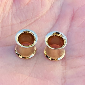 May include: Two gold-colored ear gauges with a polished finish. The gauges have a flared design with a gold rim. The tunnels are cylindrical and appear to be hollow. The gauges are shown on a light background.
