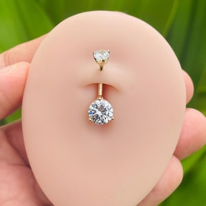 Op de afbeelding: Een gouden navelpiercing met een ronde, heldere edelsteen aan de onderkant en een kleinere ronde, heldere edelsteen aan de bovenkant.