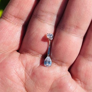 Könnte beinhalten: Ein silberner Bauchnabelpiercing mit zwei klaren, funkelnden Zirkonia-Steinen. Der obere Stein ist herzförmig, der untere tropfenförmig. Der Schmuck wird in einer Hand gehalten.