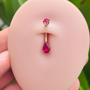 May include: A gold belly button ring with two red gemstone accents. The top gem is round and the bottom gem is teardrop shaped.