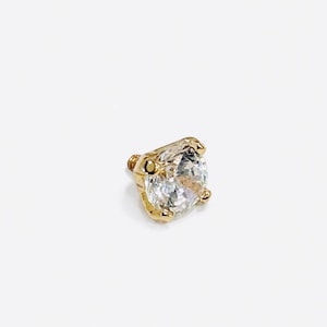 May include: A gold-colored stud earring with a large, clear, round gemstone. The gemstone is held in place by four prongs. The earring has a threaded post for secure fastening. The design is simple and elegant.