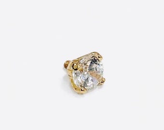May include: A gold-colored stud earring with a large, clear, round gemstone. The gemstone is held in place by four prongs. The earring has a threaded post for secure fastening. The design is simple and elegant.