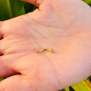 以下が含まれることがあります： 両端に小さな透明な宝石が付いた、湾曲した金色の眉ピアス。ジュエリーは開いた手のひらに保持され、背景はぼやけた緑色です。