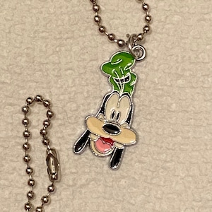 May include: A silver-colored ball chain necklace with a pendant featuring a cartoon character. The pendant is a colorful enamel design of a dog-like character with a green hat, black ears, and a pink tongue.