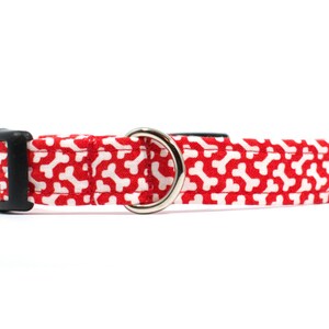 May include: Red dog collar with a white bone pattern. The collar has a black buckle and a silver D-ring.