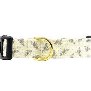 May include: A white dog collar with a gold metal buckle and a pattern of small bees. The collar is made of fabric and has a black plastic buckle.