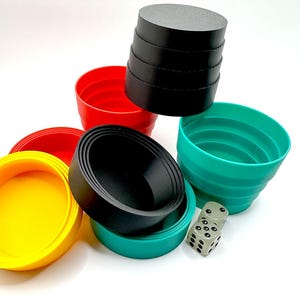 May include: A collection of colorful, stackable cups in red, yellow, teal, and black. A stack of black discs and a six-sided die are also present. The items are arranged on a white surface, suggesting a game or educational toy.