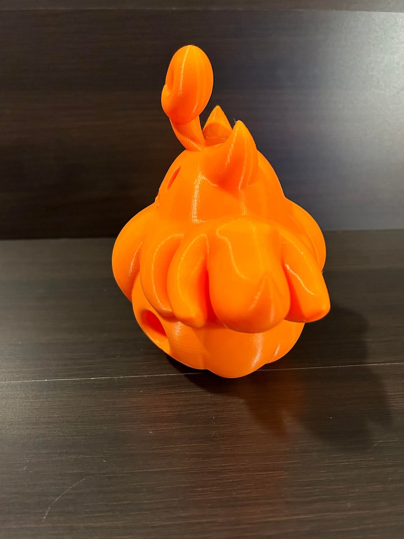 May include: An orange 3D-printed decorative object. The object has a bulbous body with petal-like structures around the base. A curved, antenna-like element extends from the top. The object is a solid, bright orange color.
