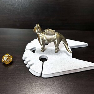 May include: A gold-colored 3D printed figurine of a cat wearing a backpack, standing on a white cat-shaped base. A gold-colored 20-sided die is in the foreground.