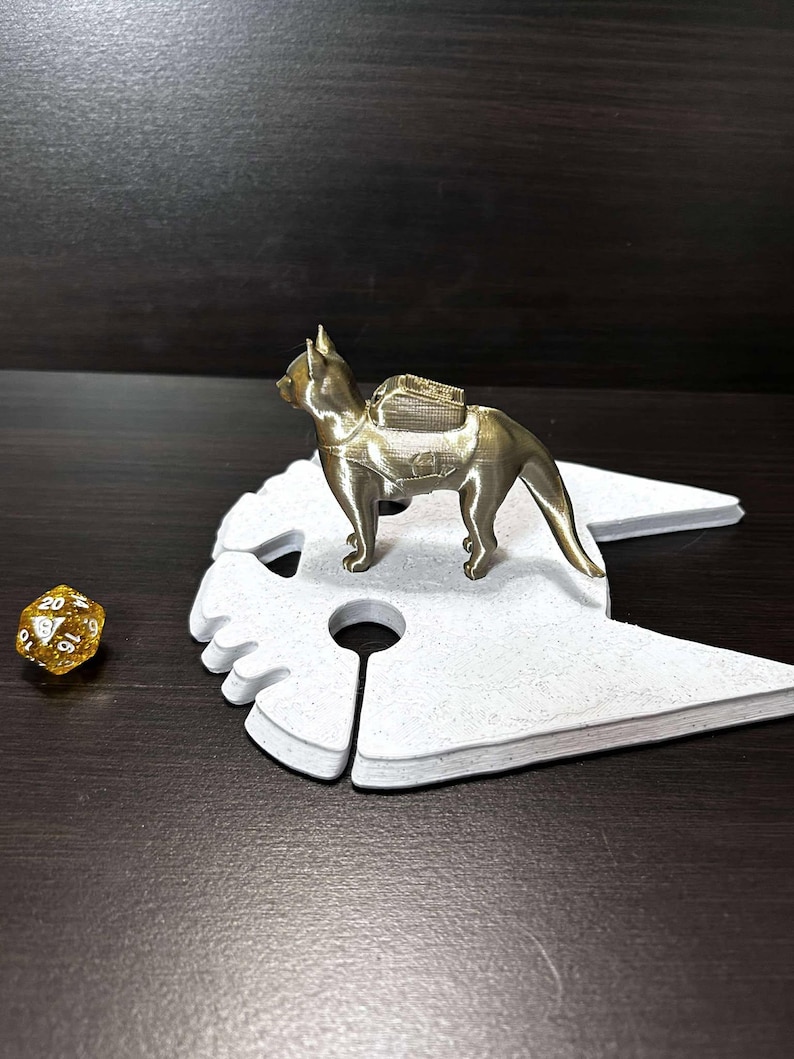 May include: A gold-colored 3D printed figurine of a cat wearing a backpack, standing on a white cat-shaped base. A gold-colored 20-sided die is in the foreground.