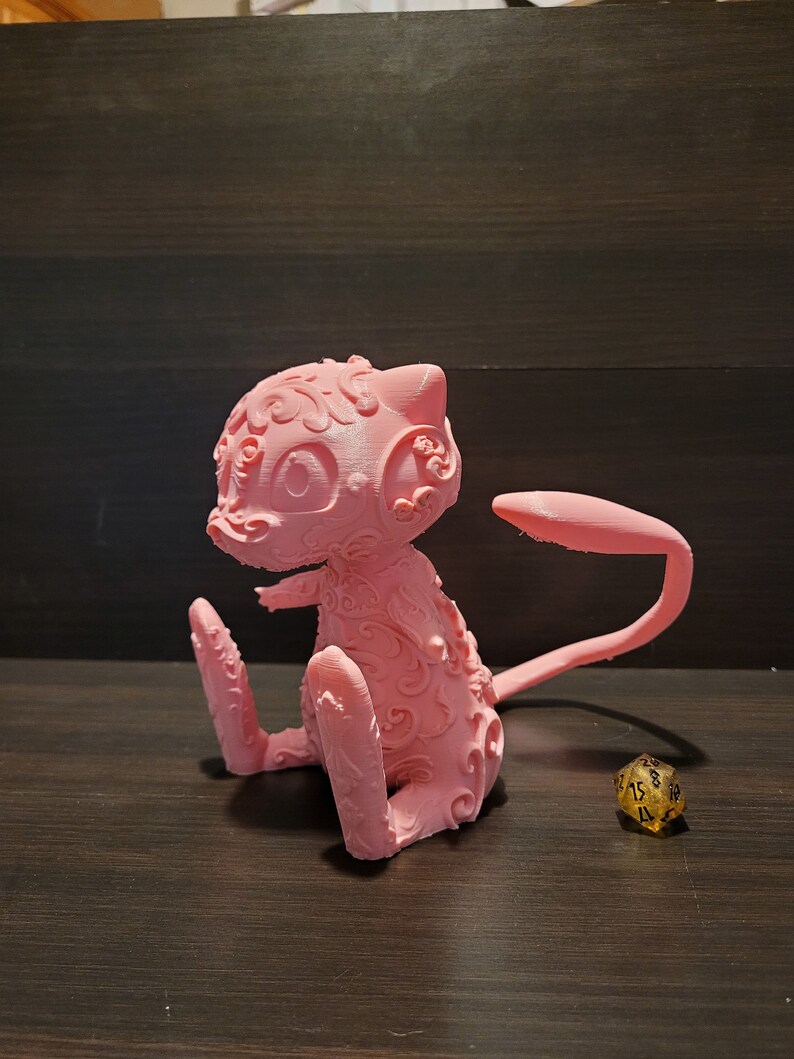 Op de afbeelding: Een roze 3D-geprint beeldje van een Pokemon-personage, met ingewikkelde details en een gekrulde staart. Het beeldje staat op een houten oppervlak.