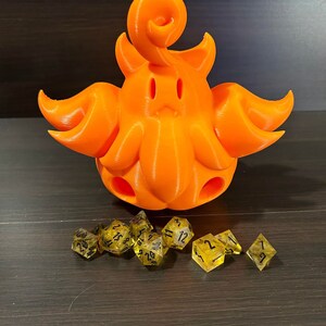 May include: An orange 3D-printed creature with a curly top and tentacle-like appendages. Several yellow and black dice are arranged in front of the creature, suitable for tabletop gaming.