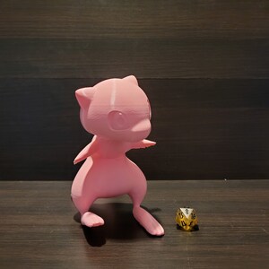 Op de afbeelding: Een roze 3D-geprint beeldje van een Pokemon-personage, mogelijk een Mewtwo, staand op een houten oppervlak met een gouden 20-zijdige dobbelsteen ervoor.
