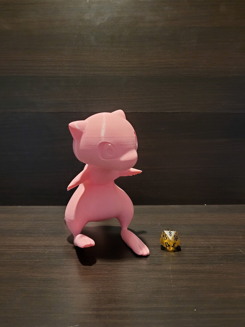 Op de afbeelding: Een roze 3D-geprint beeldje van een Pokemon-personage, mogelijk een Mewtwo, staand op een houten oppervlak met een gouden 20-zijdige dobbelsteen ervoor.