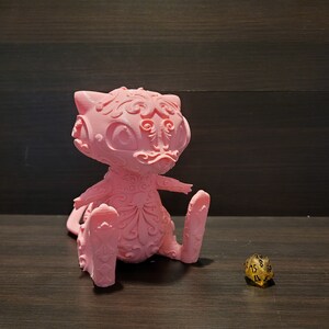 Op de afbeelding: Een roze 3D-geprint beeldje van een gestileerde kat met ingewikkelde details. De kat zit met zijn rechterpoot omhoog en een gouden 20-zijdige dobbelsteen bevindt zich op de voorgrond.