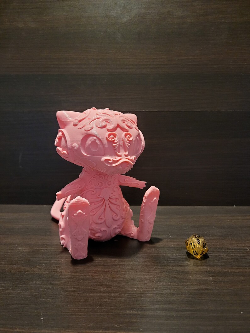 Op de afbeelding: Een roze 3D-geprint beeldje van een gestileerde kat met ingewikkelde details. De kat zit met zijn rechterpoot omhoog en een gouden 20-zijdige dobbelsteen bevindt zich op de voorgrond.