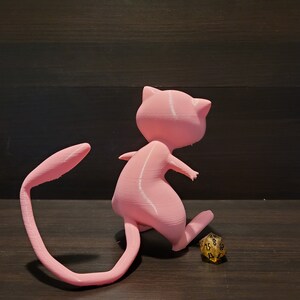Op de afbeelding: Een roze 3D-geprint beeldje van een Pok&eacute;mon-personage, mogelijk een Eevee, met een gekrulde staart. Het beeldje staat op een houten oppervlak naast een gouden dobbelsteen.