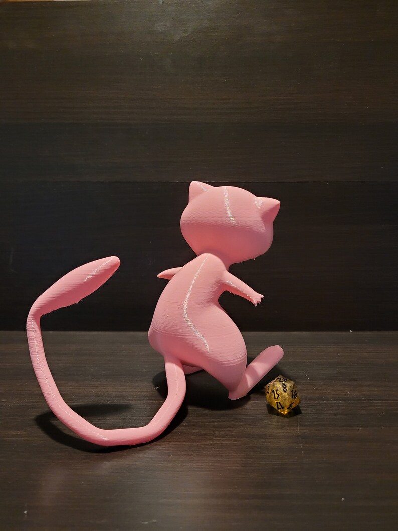 Op de afbeelding: Een roze 3D-geprint beeldje van een Pok&eacute;mon-personage, mogelijk een Eevee, met een gekrulde staart. Het beeldje staat op een houten oppervlak naast een gouden dobbelsteen.
