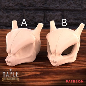 Puede incluir: Dos máscaras de calavera de plástico blanco impresas en 3D, etiquetadas como A y B, sobre una superficie de madera. Las máscaras son detalladas y se asemejan a un personaje de Pokemon.