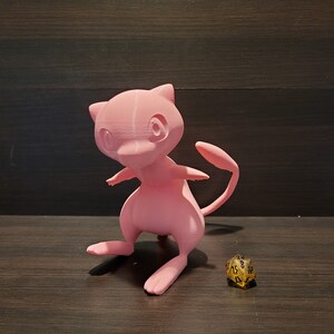 Op de afbeelding: Een roze 3D-geprint beeldje van een Pokemon-personage, mogelijk een Mewtwo, dat op een houten oppervlak staat naast een gouden 20-zijdige dobbelsteen.