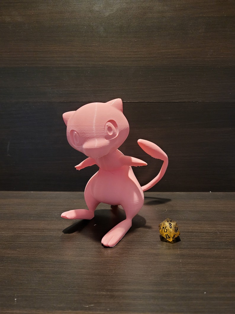 Op de afbeelding: Een roze 3D-geprint beeldje van een Pokemon-personage, mogelijk een Mewtwo, dat op een houten oppervlak staat naast een gouden 20-zijdige dobbelsteen.