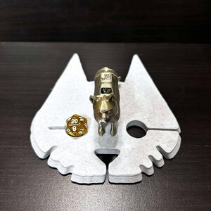 May include: A white, 3D printed dice tray shaped like a cat's head with a gold metal cat figurine standing on top. A gold metal 20-sided die is resting on the tray.