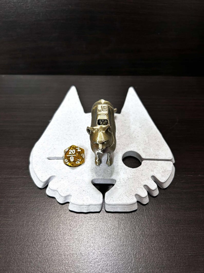 May include: A white, 3D printed dice tray shaped like a cat's head with a gold metal cat figurine standing on top. A gold metal 20-sided die is resting on the tray.