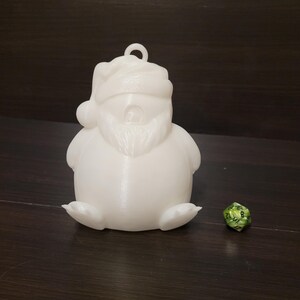 May include: White 3D printed Santa Claus ornament with a beard and a hat. The ornament is sitting on a dark brown surface next to a green and black dice.