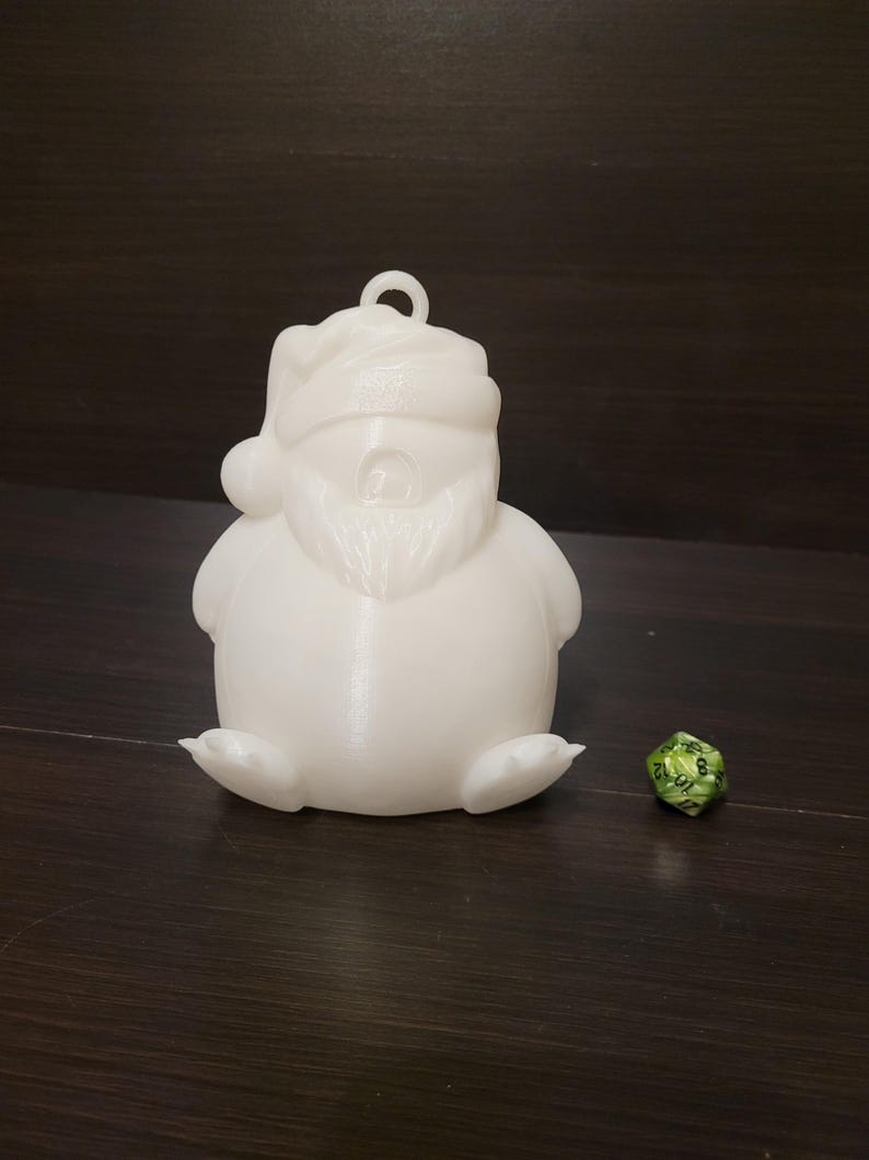 May include: White 3D printed Santa Claus ornament with a beard and a hat. The ornament is sitting on a dark brown surface next to a green and black dice.