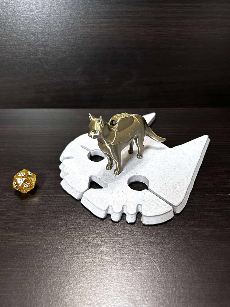 May include: A gold-colored metal cat figurine with a saddle on its back, standing on a white, textured, cat-shaped base. A gold-colored twenty-sided die is in the foreground.