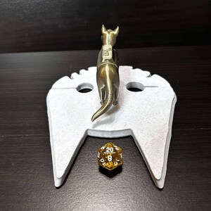 May include: A white 3D printed tabletop game accessory shaped like a spaceship with a gold-colored miniature dog figure standing on top. A gold-colored 20-sided die is on the spaceship's landing pad.