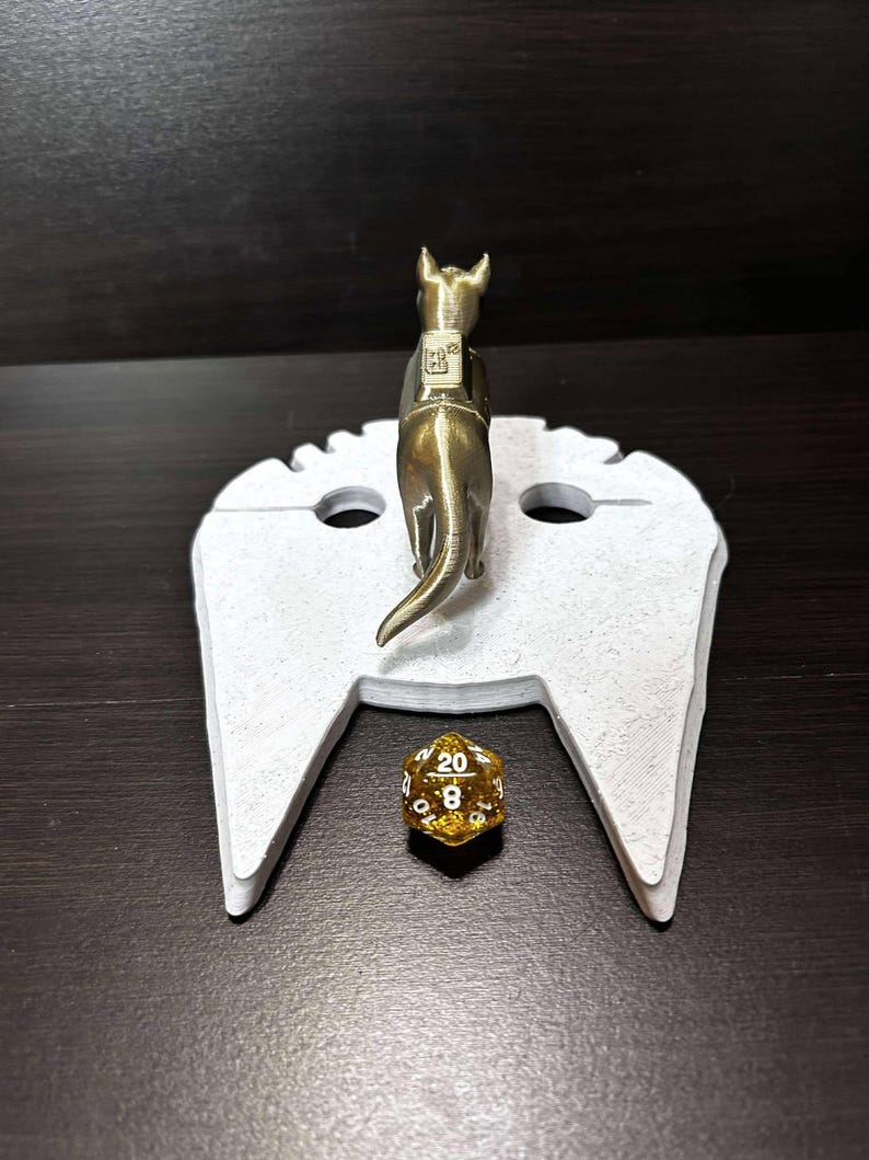 May include: A white 3D printed tabletop game accessory shaped like a spaceship with a gold-colored miniature dog figure standing on top. A gold-colored 20-sided die is on the spaceship's landing pad.