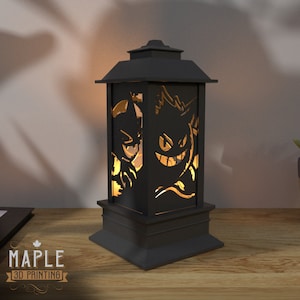 May include: A black lantern with a cutout design of two ghostly figures. The lantern is lit from within, casting a warm glow.
